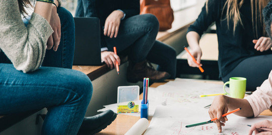 Vier Menschen sitzen an einem Tisch, die Gesichter sind nicht im Bild. Sie halten Stifte in der Hand und sehen nachdenkend aus. Eine Hand schreibt etwas auf das Plakat auf dem Tisch, welches sehr unstrukturiert gestaltet ist. Auf dem Tisch sind noch mehr Stifte und eine Tasse. Auf der Sitzbank erkennt man einen geöffneten Laptop und einen Rucksack im Hintergrund.