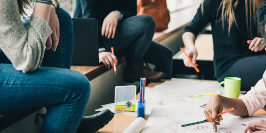 Vier Menschen sitzen an einem Tisch, die Gesichter sind nicht im Bild. Sie halten Stifte in der Hand und sehen nachdenkend aus. Eine Hand schreibt etwas auf das Plakat auf dem Tisch, welches sehr unstrukturiert gestaltet ist. Auf dem Tisch sind noch mehr Stifte und eine Tasse. Auf der Sitzbank erkennt man einen geöffneten Laptop und einen Rucksack im Hintergrund.