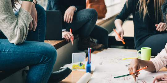 Four people are sitting at a desk writing something on a poster. There are also pencils and a cup on the table.