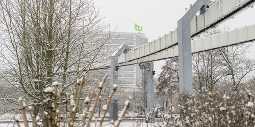 Kahle Büsche im Vordergrund, im Hintergrund H-Bahn-Strecke und das Mathematikgebäude im Schnee.